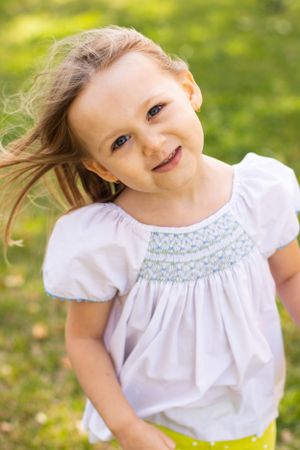 A child in a white smocked top smiles brightly against a sunlit green background.