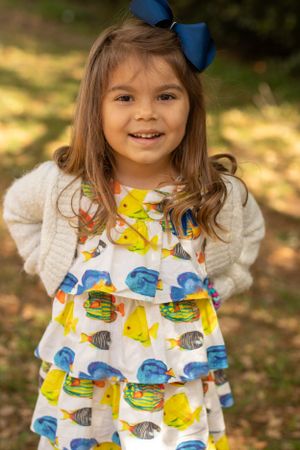 A child wearing a fish-print dress and blue bow poses with a bright smile.