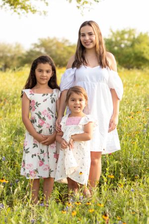 Three people in summer dresses pose together in a sunny wildflower field.