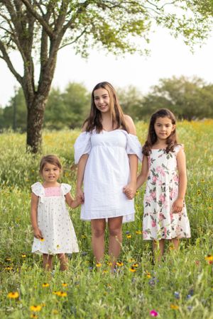 Three people in white and floral dresses stand together in a blooming wildflower field.