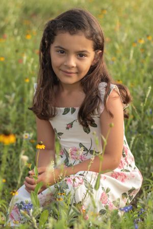 Young girl in floral dress sits among wildflowers in a sunny meadow.