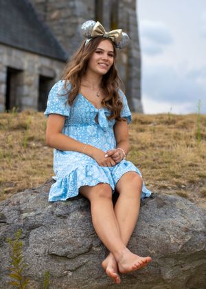 A person in a blue dress and gold mouse ears sits on a rock near a stone building.