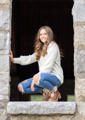 A person in a white sweater and ripped jeans poses in a stone window frame.