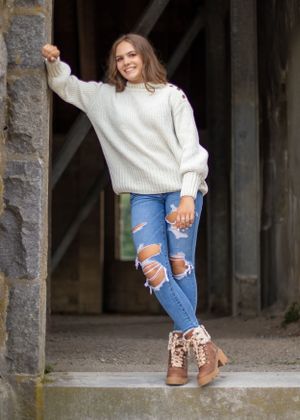 A casual fashion portrait of someone in a white sweater and ripped jeans posing against a stone pillar.