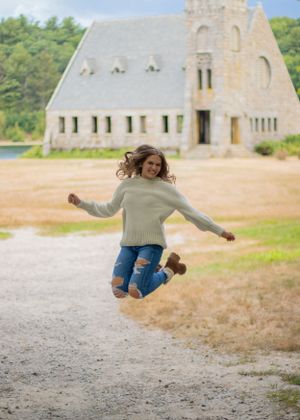 Someone in a sweater and ripped jeans jumping joyfully in front of a stone church building.
