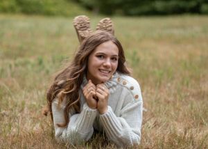 Someone with long hair lying in a grassy field wearing a white sweater and animal ear headband.