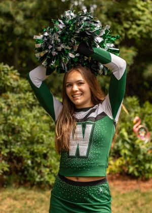 A cheerleader in green and white uniform holding pom poms overhead in an outdoor setting.