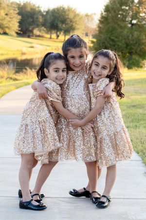 Three young children in matching sparkly gold dresses pose playfully on a pathway near a pond.