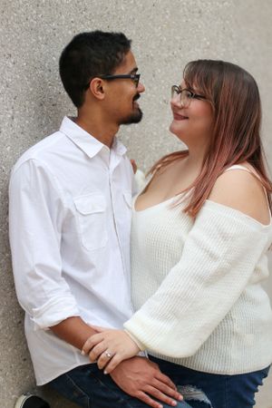 Couple in casual wear poses together against concrete wall.