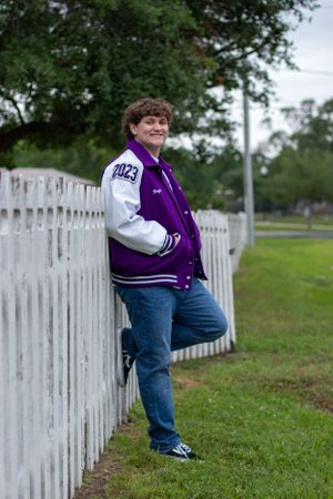 Student in purple and white varsity jacket poses by white picket fence.