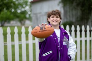 Person in purple varsity jacket holds NFL football in front of white picket fence.