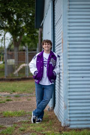 Casual pose against blue storage unit wearing purple varsity jacket and jeans.