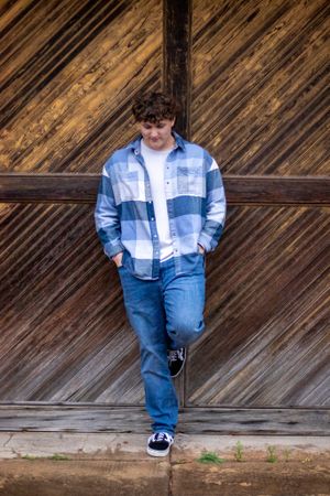 Person in plaid shirt and jeans poses against rustic wooden barn wall.