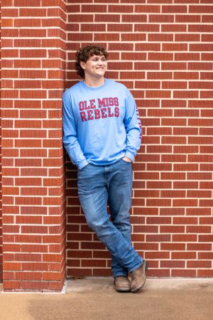 Person wearing Ole Miss Rebels sweatshirt leans against brick wall in casual denim look.