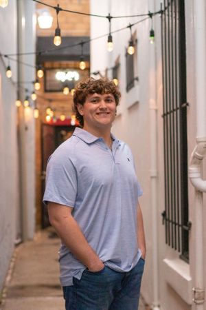 A person in a light blue polo shirt stands smiling in an alley with string lights overhead.