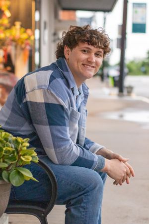 Person in plaid shirt sits on bench along storefront sidewalk with plants nearby.