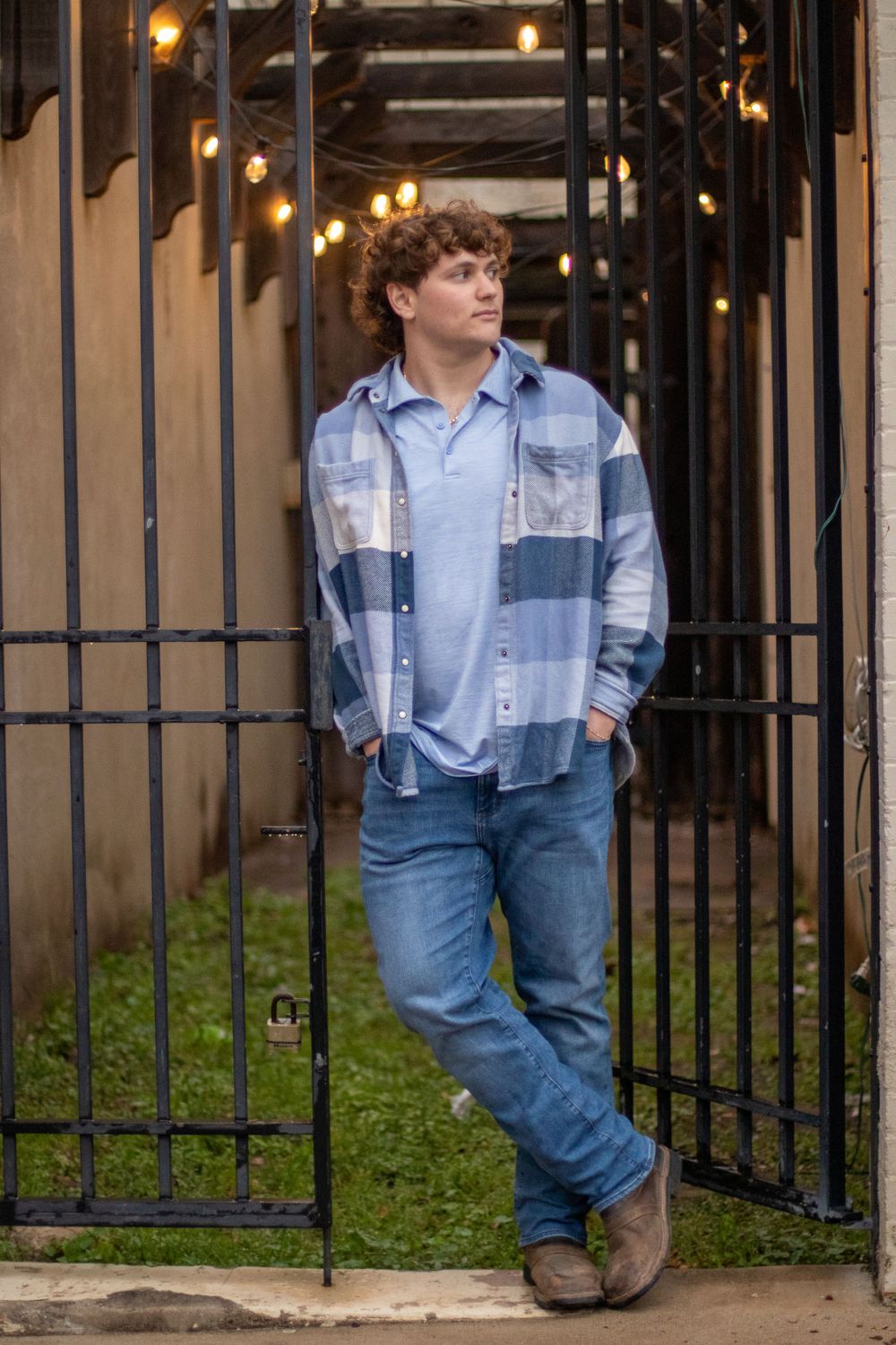 Person in a plaid blue button-down shirt and jeans poses against metal gates with string lights overhead.