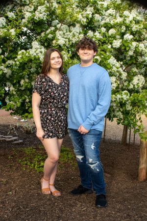 Two people stand together in front of a blooming white floral backdrop in casual spring attire.