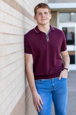 A person in a burgundy polo shirt leans against a beige wall.