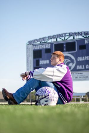 A person sits on grass near a football scoreboard wearing purple and white athletic gear.
