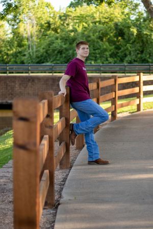 Someone in a burgundy shirt and jeans leans against a wooden railing on an outdoor path.