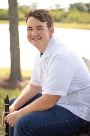 Student musician in white polo sits outdoors holding a clarinet.