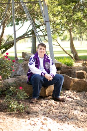 Someone sits on stone landscaping wearing a purple and white varsity jacket.
