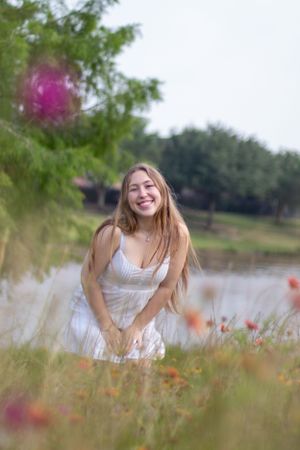 Person in white dress smiling joyfully in flowering meadow near a lake.