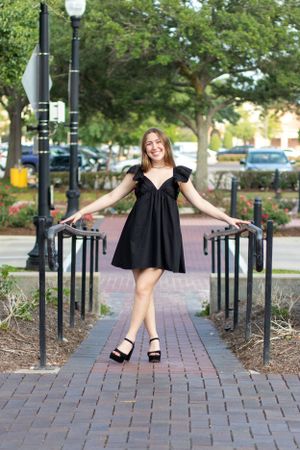 Person in black dress strikes playful pose on brick pathway between lampposts.