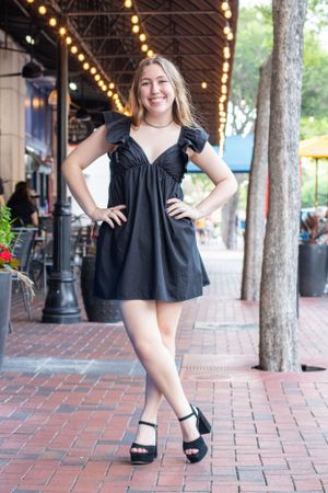 Person in black summer dress poses confidently on brick sidewalk under string lights.