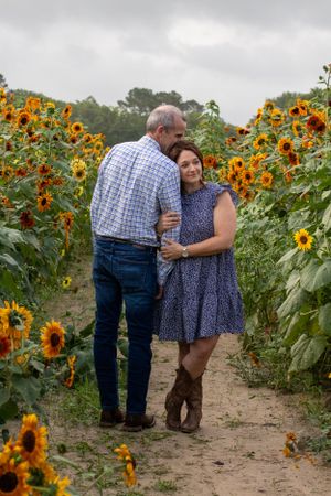 Romantic couple embracing on dirt path surrounded by towering sunflowers on cloudy day.