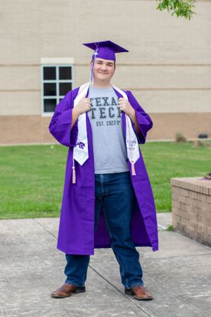 Graduate in purple cap and gown wearing a Texas Tech shirt stands proudly on campus grounds.