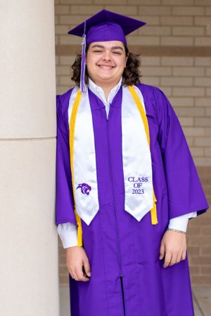 Graduate wearing purple and gold graduation regalia with Class of 2023 stole poses by brick wall.