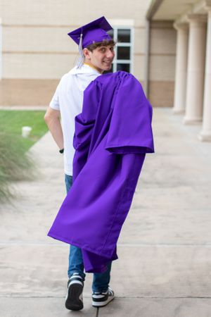 Graduate in purple regalia walks along campus pathway carrying graduation gown.