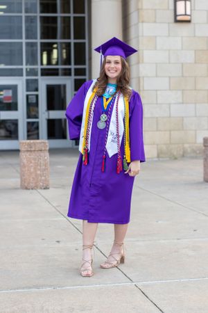 Graduate in purple graduation gown and cap poses with honors cords outside campus building.