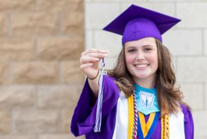 Graduate in purple cap and gown holds tassel while posing against stone wall.