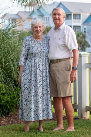 An elderly couple stands together on their lawn wearing casual summer attire near coastal beach houses.