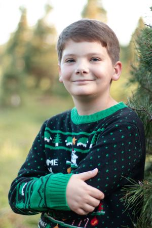 Young boy wearing festive green Christmas sweater poses near evergreen trees.