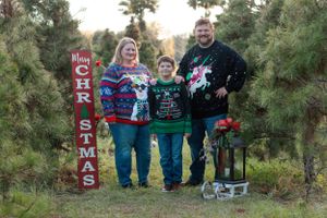 Family wearing matching Christmas sweaters poses by holiday decorations at tree farm.