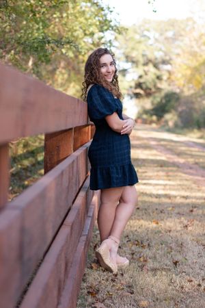 A young woman in a navy dress poses against a rustic wooden fence on an autumn path.