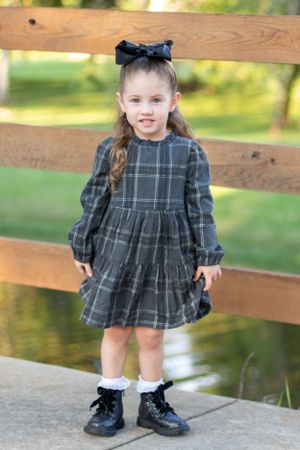 Young girl in a plaid dress and bow poses near wooden fence rails outdoors.