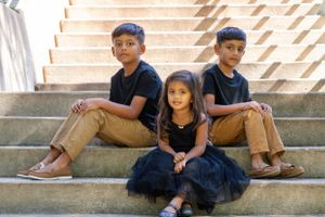 Three siblings in coordinated outfits pose casually on concrete steps.