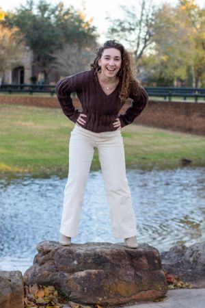 An energetic pose on a rock by a pond wearing white pants and a brown sweater.