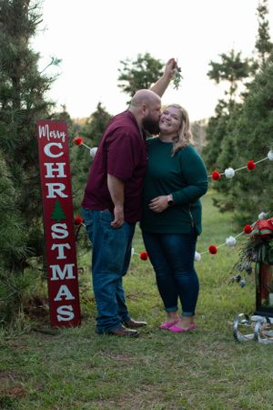 A couple shares an intimate moment by a rustic wooden 'Merry Christmas' sign among evergreen trees.