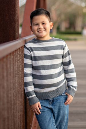 Young boy in striped sweater smiling by wooden railing outdoors.