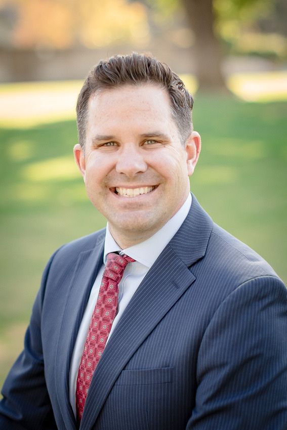 Professional headshot of person in navy pinstripe suit with red tie against blurred outdoor background.