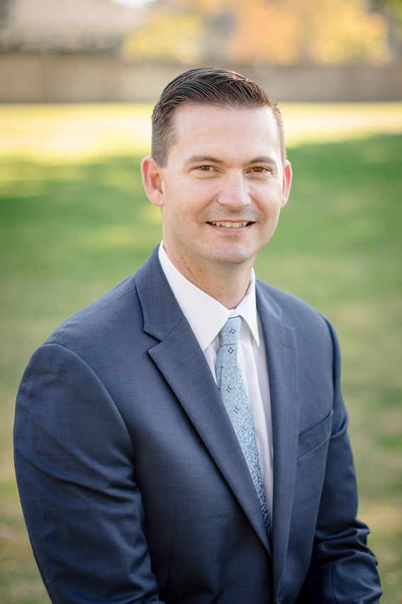 Professional headshot of person wearing navy suit and light blue tie outdoors.