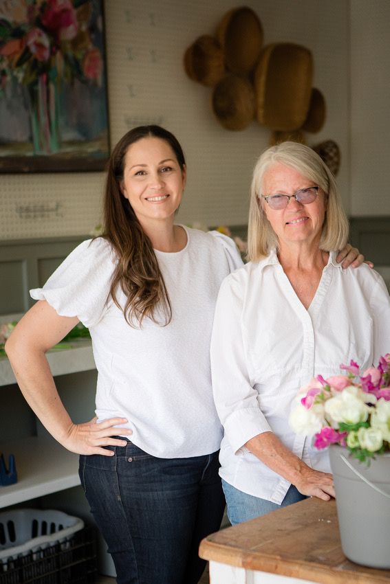 Two people in white shirts pose together in a kitchen with floral artwork and decorative plates on the wall.