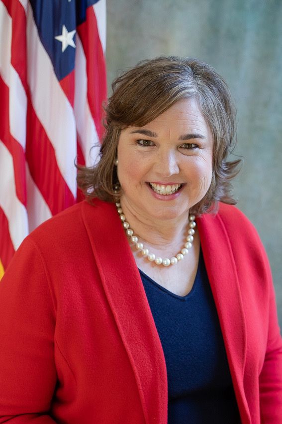 Professional headshot of person wearing red blazer, pearl necklace and navy top against American flag.