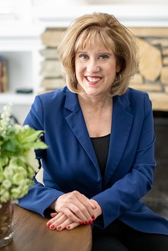 Professional portrait of person in navy blazer seated at desk with greenery in background.
